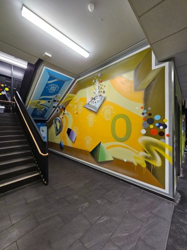 Innenraumgestaltung Treppenhaus – Magie des Lernens 3D-Graffiti-Kunst an einer gelben Innenwand im Treppenhaus mit geometrischen Körpern und schwebenden Icon-Elementen die Schule symbolisieren
