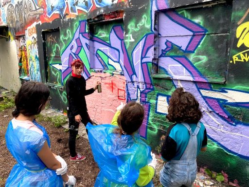 Graffiti Workshop mit Kids Kinder in blauen Regencapes und Schutzkleidung stehen vor einer bunten Graffitiwand im Freien.