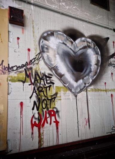 Make Art Not War Ein plastisch gesprühter Herzballon über dem Schriftzug "Make Art Not War" mit Einschusslöchern an einer Betonwand.