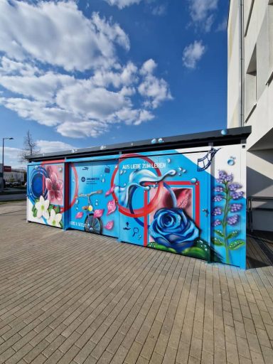 Funktionsgebäude Johanniter – „Aus Liebe zum Leben“ Bunt gestalteter Funktionsbau der Johanniter mit Graffitikunst aus Rosen, Blumen, Herz-Motiv und Schriftzug Aus Liebe zum Leben.