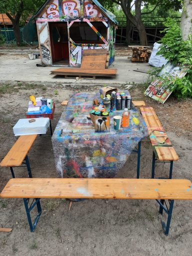 Outdoor Workshop Arbeitsplatz Eine Bierzeltgarnitur im Freien mit einem bunt beklecksten Arbeitstisch voller Malutensilien und Sprühdosen.