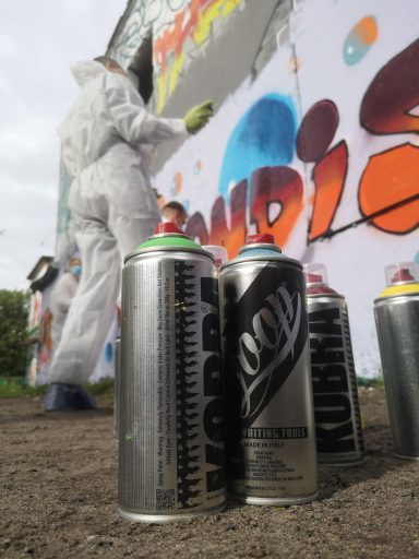 Firmenteam at Work - Sprühdosen Nahaufnahme Nahaufnahme von verschiedenen Graffitisprühdosen auf einem Tisch mit verschwommenem Hintergrund einer sprayenden Person.