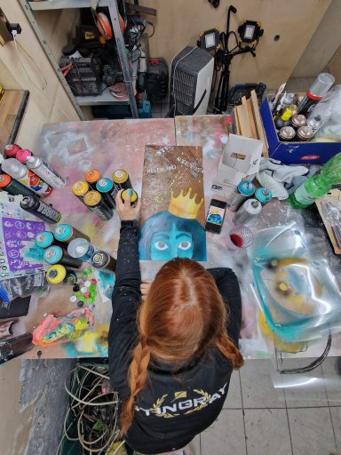 Künstlerin bei der Arbeit Blick von oben auf eine rothaarige Person, die an einem kreativen Tisch mit Farben und Pinseln malt.