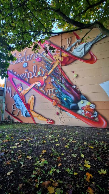 Fassadenkunst Sporthalle – der sportliche Funke Großflächiges Graffiti-Wandbild an der Außenfassade einer Sporthalle mit zwei dynamischen Athleten in Sportkleidung und einem Ball.