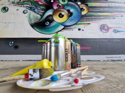 Kunst-Equipment und Augen-Mural Sprühdosen, Mischpalette und Pinsel liegen im Vordergrund vor einem Graffiti mit großen Augen.