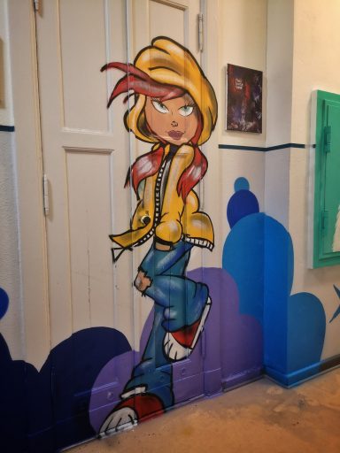 Street Kids Club - Gestaltung Eine Graffiti-Malerei einer stilisierten Figur mit einer gelben Kapuzenjacke und blauer Hose auf einer weißen Flurwand.