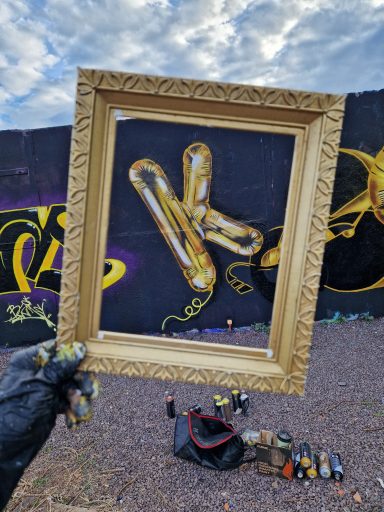 Ballonletter catched Eine Hand hält einen goldenen Bilderrahmen vor ein Graffiti mit dem Buchstaben K in Ballonoptik.