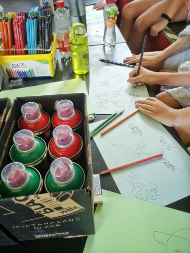 Kinder skizzieren und bereiten vor Hände von Kursteilnehmern beim Zeichnen mit Buntstiften auf Papier neben einer Kiste voller Sprühdosen.