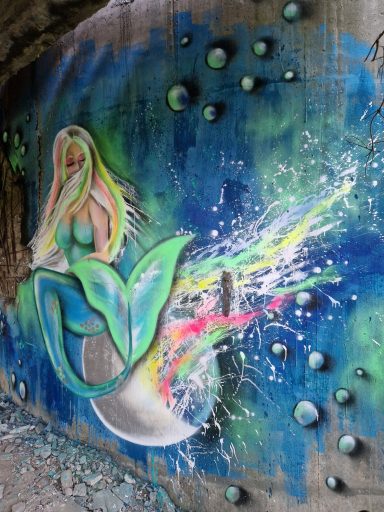 Meerjungfrau in Neonfarben Wandgemälde einer Meerjungfrau in Blau- und Grüntönen mit bunten Sprühfarbeneffekten.