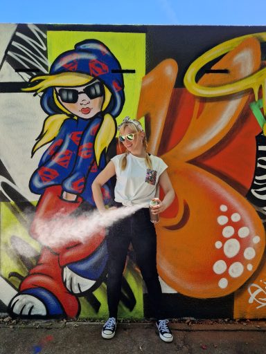 Künstlerin vor Graffiti-Wand Eine Frau steht mit einer Sprühdose in der Hand vor einer bunt bemalten Graffiti-Wand.