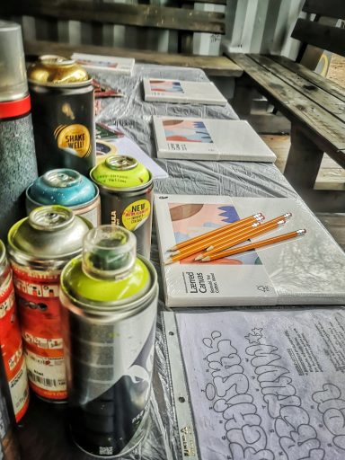 Skizzen und Sprühdosen Set Sprühdosen, ein Stapel Bleistifte und vorbereitete Skizzenpapiere auf einem Tisch im Freien.