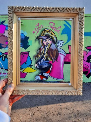 Graffiti Kunst im Bilderrahmen Hand hält ein kleines eingerahmtes Bild einer Comic-Figur vor einer bunt besprühten Wand.
